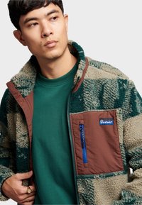 Fleece jacket in green and tan with a patterned texture, featuring a brown chest pocket and blue zipper. Layered over a green shirt.
