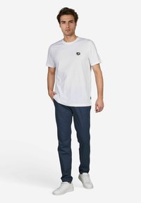 Weißes Baumwoll-T-Shirt mit einem Sonnenstrahl-Design, kombiniert mit navyblauen, taillierten Hosen und weißen Sneakers, vor einem schlichten Hintergrund.