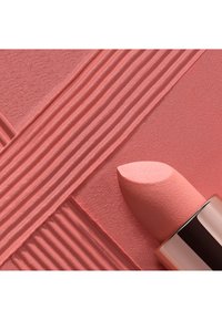 Lichtroze lipstick met een zijdezachte textuur en een schuine punt, geplaatst op een gestructureerde roze achtergrond met verticale en horizontale lijnen.