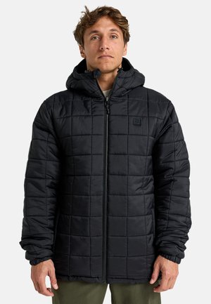 Hombre vestido con una chaqueta con capucha negra acolchada y cremallera frontal, de pie contra un fondo claro y liso.