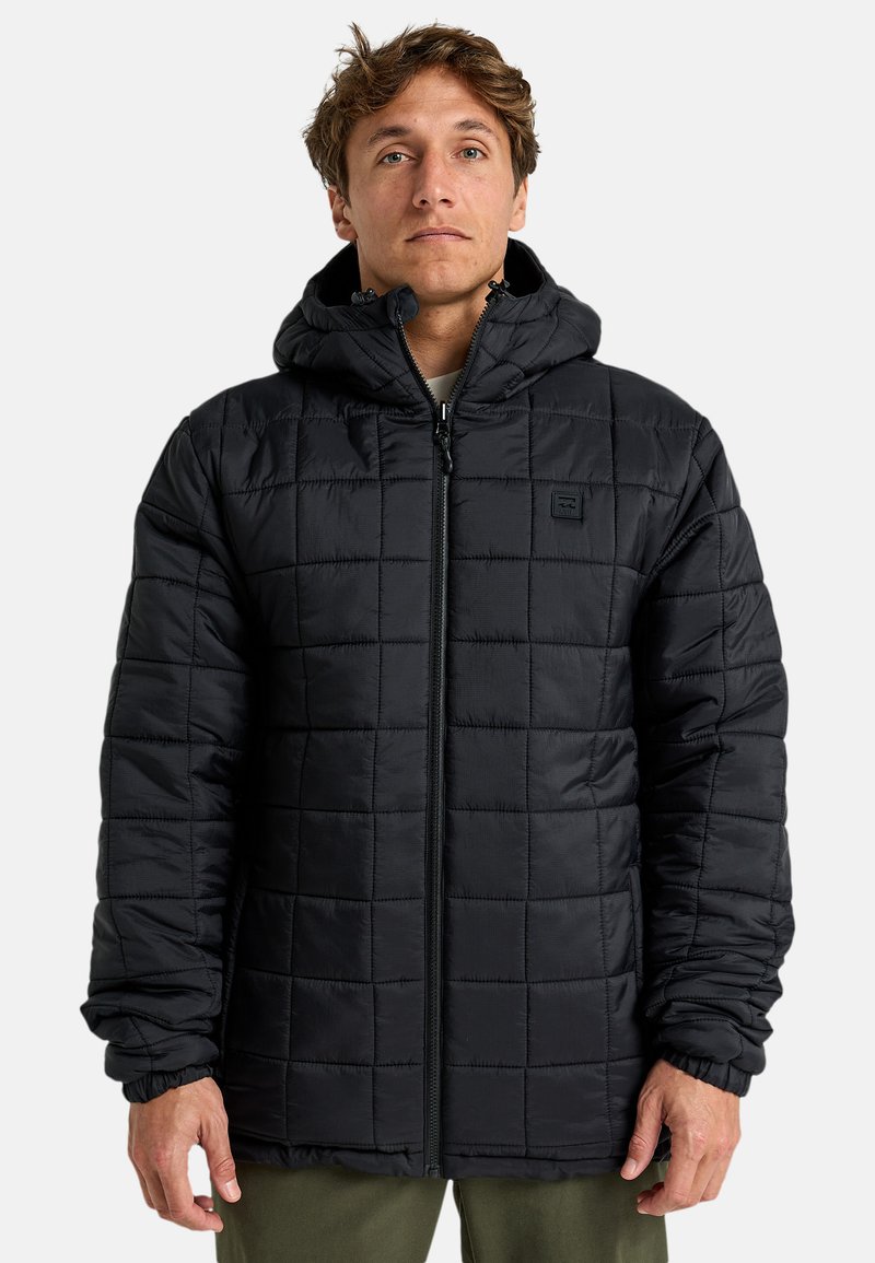 Hombre vestido con una chaqueta con capucha negra acolchada y cremallera frontal, de pie contra un fondo claro y liso.