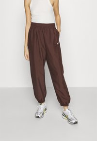 Hnědé, lehké, elastické kalhoty s manžetami a bočními kapsami, na levé straně s bílým logem Nike. Kombinované se šedými a žlutými teniskami.