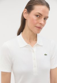 Biele rebrované polo tričko s golierom a piatimi gombíkmi, s malým zeleným krokodílom na ľavej hrudi.