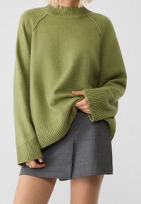 Person, der einen übergroßen olivgrünen Strickpullover mit weiten Bündchen und einem grauen, gefalteten Minirock trägt, zieht den Pullover leicht nach vorne.