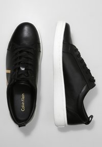 Černé kožené tenisky Calvin Klein s bílou podešví, jedna bota z pohledu shora a jedna z bočního pohledu na světlém šedém pozadí.