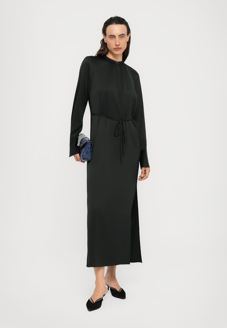 Femme portant une longue robe noire avec une taille à nouer, tenant une pochette bleue, et portant des escarpins noirs à bout pointu, se tenant contre un fond blanc.