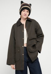 Καφέ βαμβακερό μπουφάν με γιακά από κοτλέ ύφασμα, κουμπιά τύπου snap και μπροστινή τσέπη. Φοριέται πάνω από ελαφρύ πλεκτό τοπ και συνδυάζεται με σκούρο τζιν παντελόνι.