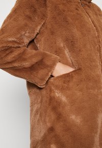 Brauner Kunstpelzmantel mit weicher Textur, ausgestattet mit einem Reißverschluss und Seitentaschen. Das Design hat eine lockere Passform und eine glatte Oberfläche.