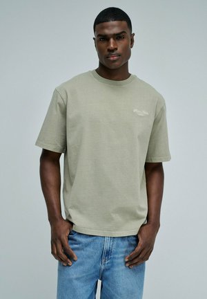 Jeune homme portant un t-shirt à manches courtes vert olive clair et un jean bleu, debout les mains près des poches devant un fond uni.