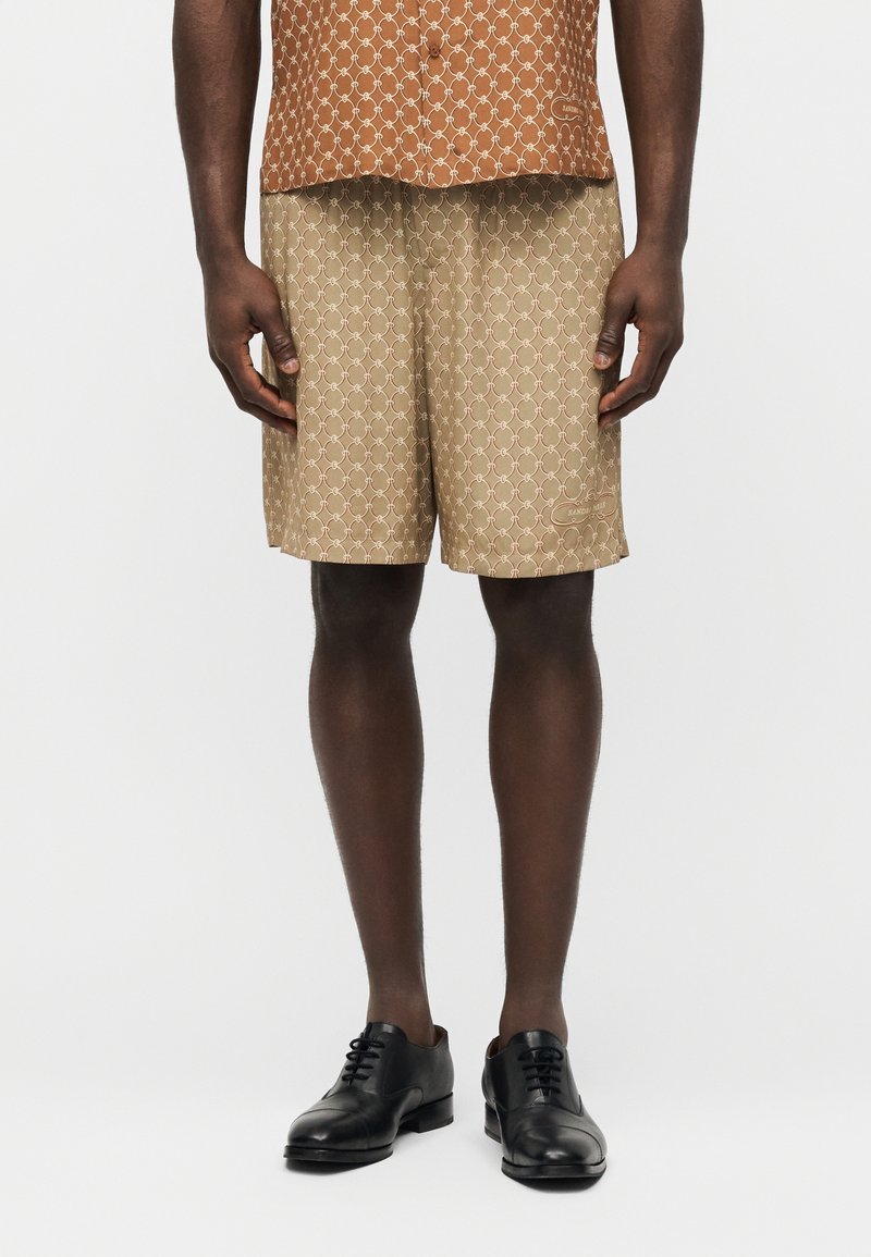 Mann trägt beige Shorts mit Muster und ein orangefarbenes Hemd mit Muster, kombiniert mit schwarzen Lederschuhen, steht vor einem schlichten Hintergrund.