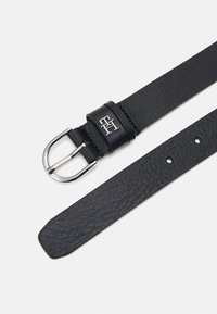 Curea din piele neagră cu o suprafață texturată, cataramă din metal argintiu și logo embossat pe curea. Dispune de mai multe ajustări prin găuri pentru dimensiune.