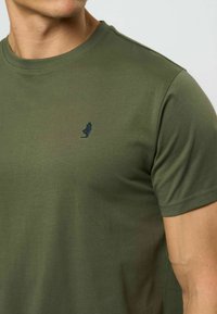 Vyras dėvi alyvuogių žalios spalvos trumpomis rankovėmis marškinėlį su mažu tamsiu siuvinėtu figūros piešiniu kairėje krūtinės pusėje.