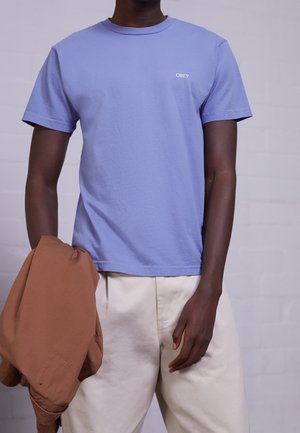 Lichtpaars katoenen T-shirt met een ronde hals en korte mouwen, met een klein wit "OBEY"-logo op de borst. Model houdt een bruine jas vast.