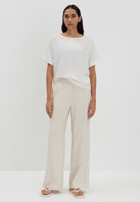 Haut blanc à manches courtes avec un col rond, associé à un pantalon beige ample. La tenue est complétée par des tongs blanches. Textures lisses.