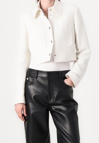 Veste blanche raccourcie avec un tissage texturé, dotée d'un col et de deux poches avant, associée à un pantalon en cuir noir.