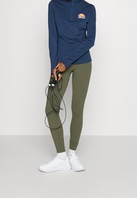 Marinblå långärmad tröja med dragkedja och logotyp, kombinerad med olivgröna leggings och vita sneakers; hållande ett svart hopprep.
