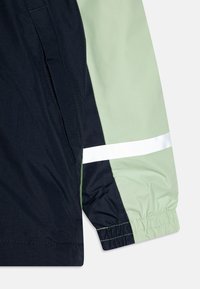 Veste coupe-vent avec un design en coloris bloc bleu marine et vert clair, dotée d'une bande réfléchissante blanche et de poignets élastiques.