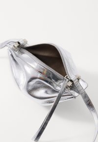 Borsa a tracolla metallica argento con superficie lucida, chiusura con zip e interno texturizzato. Presenta una tracolla staccabile e un design angolare.