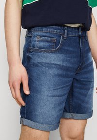 Hombre vestido con pantalones cortos de mezclilla oscuros y enrollados, y una camiseta verde y azul marino, mostrando manos y muslos superiores.