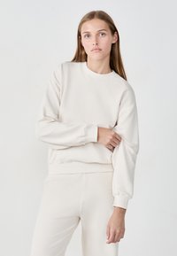 Sudadera de color beige claro hecha de una tela suave, con cuello redondo, mangas largas y puños acanalados. Diseño simple sin estampados.