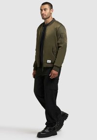 Olive green bomber jacket z zamkiem błyskawicznym, ściągaczami przy mankietach i bocznymi kieszeniami. Warstwowa konstrukcja z naszywką po lewej stronie oraz czarne spodnie cargo.