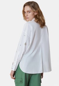 Camicia bianca a maniche lunghe con hardware decorativo sulle maniche, vestibilità rilassata e orlo arrotondato. Indossata con pantaloni cargo verdi.