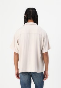 Camicia corta beige chiaro con un taglio rilassato, caratterizzata da un colletto semplice, una texture liscia e un dettaglio sul retro. Abbinata a jeans blu.