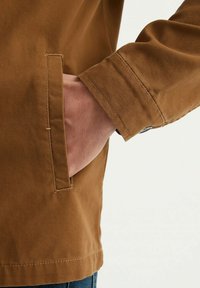 Veste marron texturée avec une poche latérale ; les coutures visibles et le détail des poignets soulignent le design. Coupe décontractée et pratique.