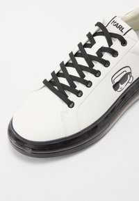 White sneaker with black laces, featuring a smooth texture and a transparent black sole. A monochrome graphic is printed on the side.