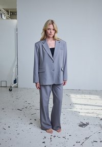 Femme blonde debout pieds nus dans un costume gris surdimensionné avec un haut noir dans une pièce lumineuse et industrielle avec un sol blanc moucheté.