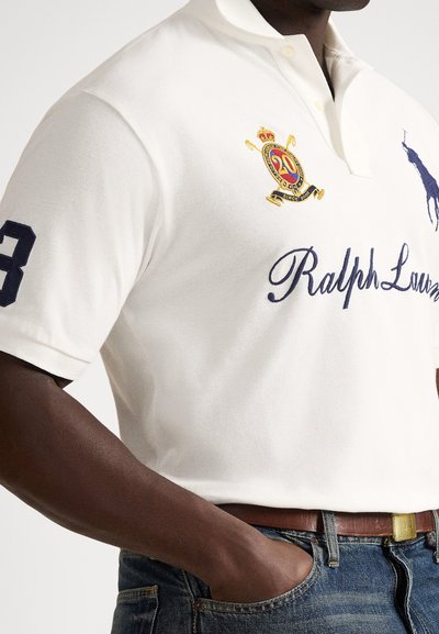 Bela polo majica Ralph Lauren z vezenim logotipom, številko 3 na rokavu, nosena zatlačena v modre kavbojke s rjavim usnjenim pasom.