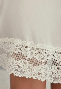 Vitt tyg med en krämfärgad spetsDetalj i fållen som har blommönster och en scalloped kant, slät textur och genomskinlig utseende.
