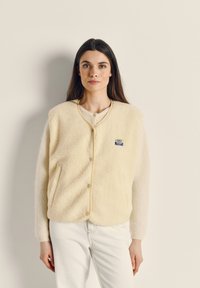 Kreminės spalvos vilnos liemenė su tekstūruota paviršiaus apdaila, spaudžiamais mygtukais, apvalia apykakle ir mažu žaliu bei mėlynu logotipo lipduku.