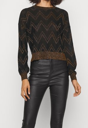 Femme portant un haut à manches longues noir et doré à motif zigzag avec des poignets métalliques et un pantalon en cuir noir taille haute.