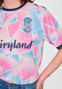 Koszulka z różowym, niebieskim i białym abstrakcyjnym wzorem, logo "Forward Madison" z flamingiem i napisem "airyland", noszona przez osobę w dżinsach.