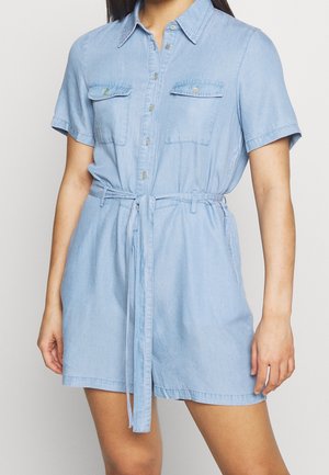 Kobieta w jasno-niebieskiej, krótkiej sukience dżinsowej z guzikiem z przodu, kieszeniami na piersiach oraz związanym paskiem w talii.