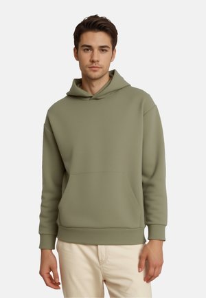 Jonge man draagt een effen olijfgroene hoodie en lichtbeige broek, staat met een neutrale uitdrukking tegen een witte achtergrond.