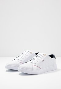 Białe casualowe sneakersy z teksturowanym wzorem, z okrągłym noskiem, białą gumową podeszwą, kontrastującymi granatowymi akcentami i złotymi oczkami.