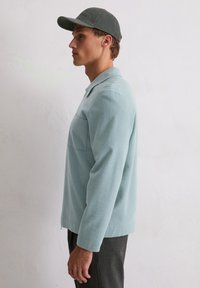 Jasnoniebieska polarowa koszula z kołnierzykiem i kieszonką na piersi, zestawiona z szarym czapką, widok z boku na neutralnym tle.