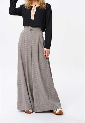 Kobieta nosząca wysokie, szerokie spodnie w kratę, czarną bluzkę z długim rękawem z beżowym obszyciem oraz białe buty sportowe z brązową podeszwą.