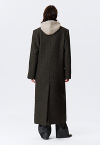 Long, dark green wool coat with a beige hood. Features a straight silhouette, textured fabric, long sleeves, and a vent at the back.