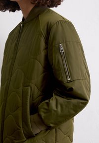 Olijfgroen gewatteerde bomberjack met elastische manchetten, zijzakken en een ritszak op de linker mouw. Glad, duurzaam stofoppervlak.