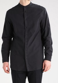 Camisa negra de manga larga con cuello mandarín, que presenta un cierre frontal de botones y una tela suave y ligera. Sin estampados ni adornos.