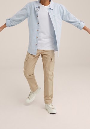 Jeune personne portant une chemise boutonnée bleu clair par-dessus un t-shirt blanc, un pantalon cargo beige et des baskets blanches, debout les bras étendus.