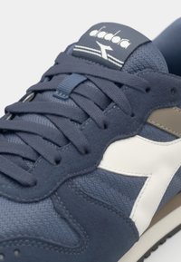 Navy blue sneaker with a textured suede and mesh upper, featuring a white logo, gray accents, and flat laces. Rubber sole with visible tread.