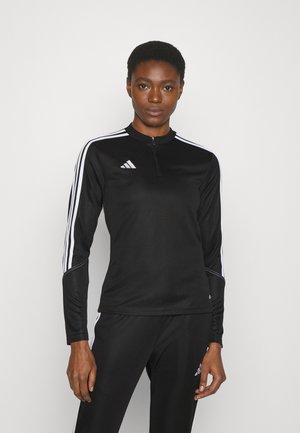 Camiseta deportiva de manga larga negra con cremallera de cuarto, diseño de tres rayas blancas en las mangas y un pequeño logo en el pecho. Textura de tela suave.