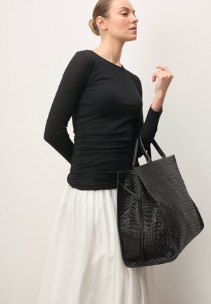 Geantă tote neagră din piele împletită cu suprafață texturată, având două mânere și un design spațios, asortată cu un top negru mulat și o fustă albă.