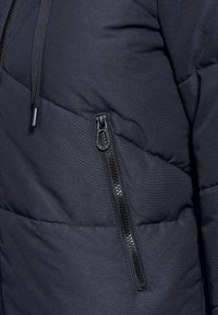 Veste rembourrée bleu marine avec un design matelassé diagonal, poche zippée noire et ajustement de capuche à cordon. Texture de tissu lisse.