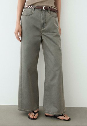 Personne portant un pantalon gris à jambes larges, une ceinture marron cloutée, un haut beige et des sandales noires à brides, debout devant un fond clair.