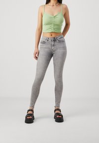 Ljusgrön avkortad topp med rynkning, kombinerad med grå tajta jeans. Modellen bär svarta platåsandaler. Neutral bakgrund.